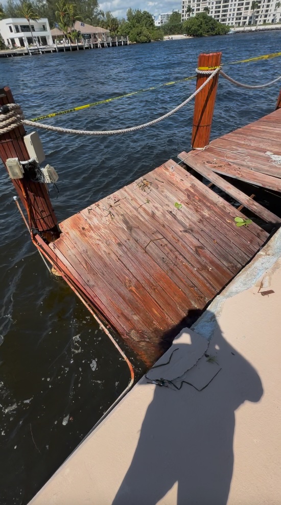 Collapsed wooden dock section dropping toward the water, with caution tape stretched across the canal